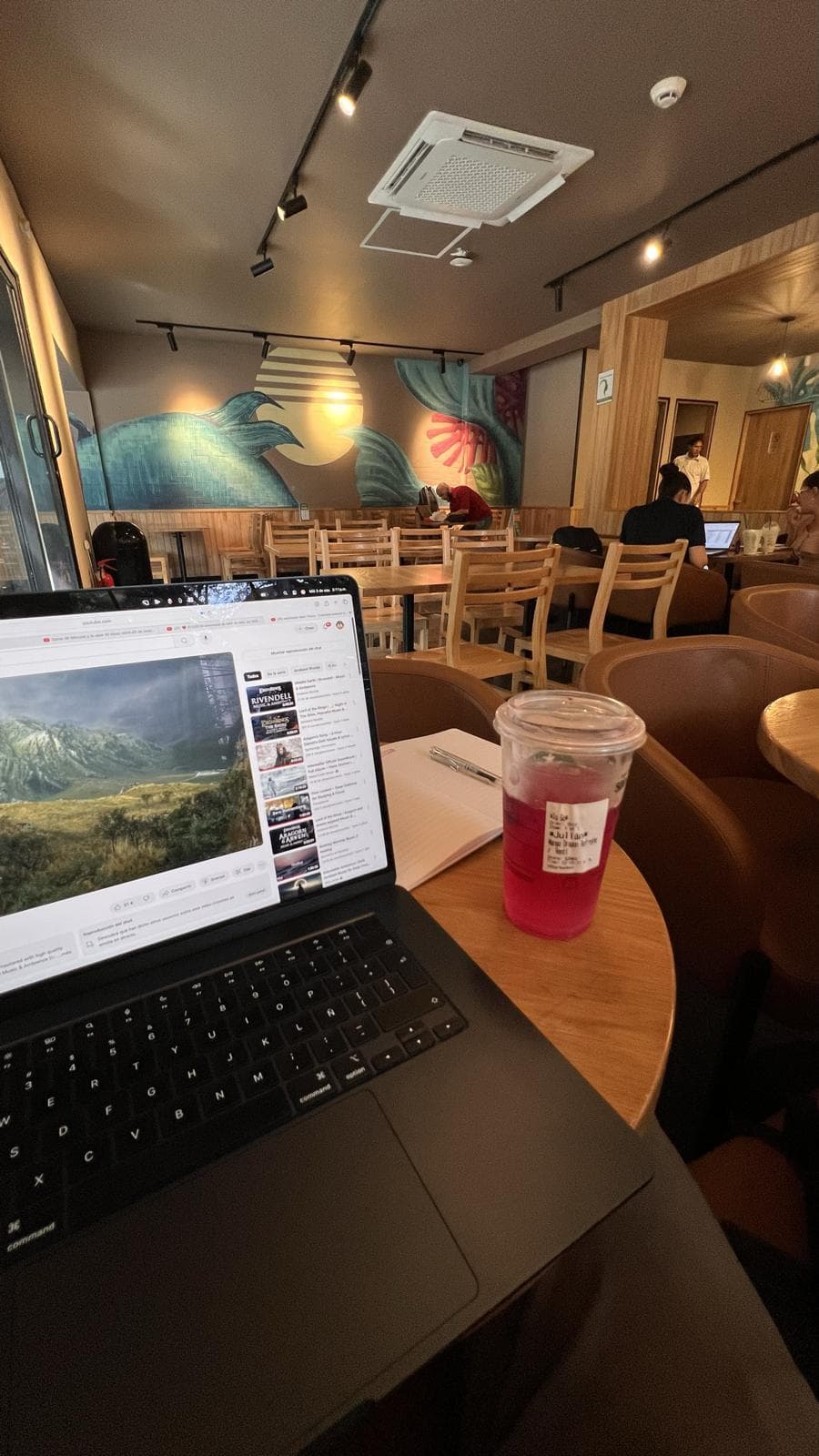 Un portátil abierto sobre una mesa redonda de madera en un café, junto a una libreta con un bolígrafo y una bebida roja con hielo. Al fondo, sillas de madera, personas trabajando y un mural con un sol y hojas tropicales.